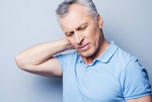 i need a massage. frustrated senior man in t-shirt holding hand on his neck while standing against grey background