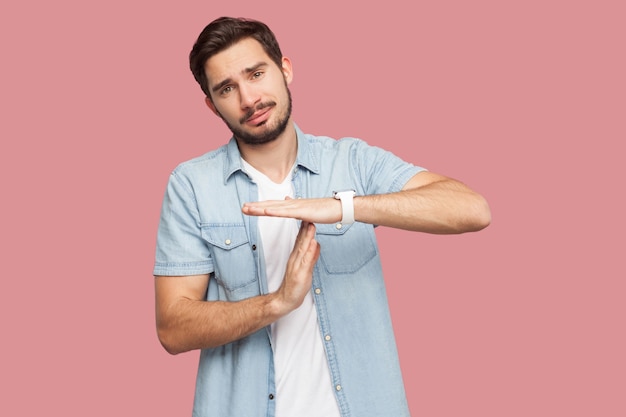 I need more time. Portrait of worry handsome bearded young man in blue casual style shirt standing with timeout gesture and begging for more time. indoor studio shot, isolated on pink background.