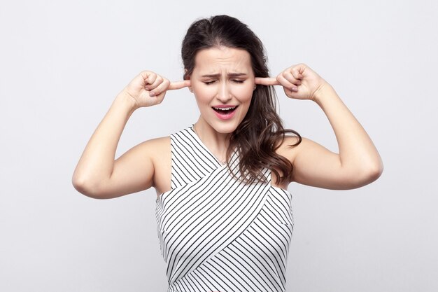 I don't want to here this. Portrait of unhappy beautiful young brunette woman with makeup and striped dress standing and holding fingers on her ear. indoor studio shot, isolated on grey background.