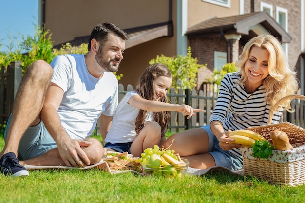 I want it. Lovely little girl sitting in between her parents on picnic and pointing at bananas, asking her mother to give them to her