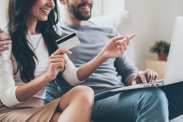 I want this! Close-up of beautiful young woman holding credit card and pointing laptop with smile while sitting together with her husband on the couch