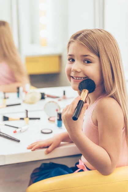 I will be ready in five minutes! Cheerful little girl applying make-up and looking at camera with smile whilesitting at the dressing table