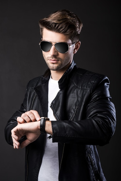 I will be in time. Handsome young man in sunglasses watching time while standing against black background