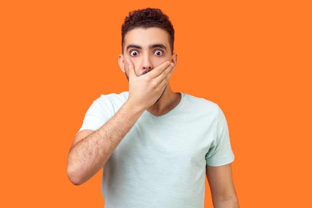 I will not say Portrait of scared bullied man in white tshirt covering mouth with hand looking with frightened shocked eyes keeping secret taboo indoor studio shot isolated on orange background