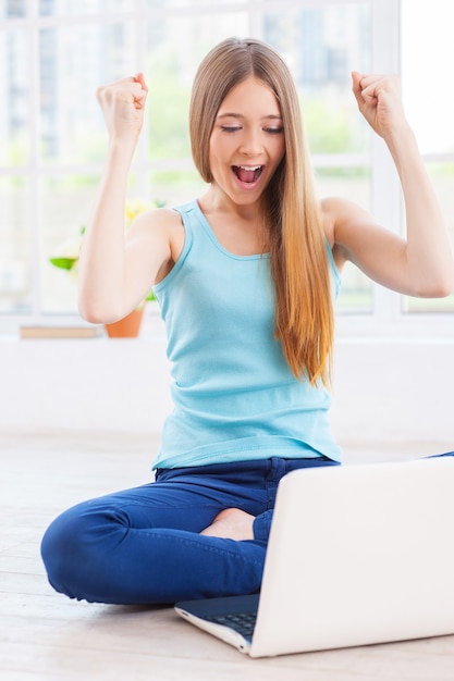 I won! Cheerful teenage girl looking at laptop and keeping arms raised while sitting on the floor at her apartment