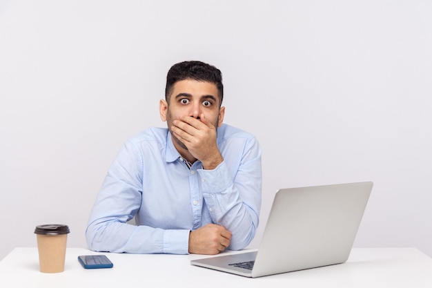 I won't tell anything. Scared man employee sitting office workplace with laptop on desk, covering mouth looking with terrified expression, knows scary secret. studio shot isolated on white background