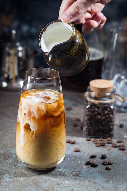 石の背景にクリームとコーヒー豆を注ぐ背の高いグラスでアイスコーヒーの冷たい夏の飲み物。