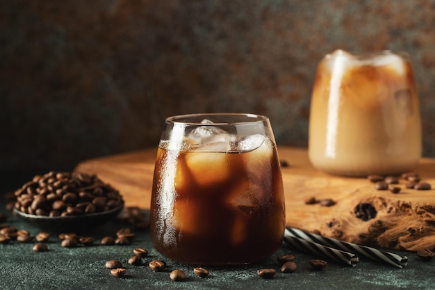 Ice coffee in a tall glass with cream poured over ice cubes and beans on a dark concrete table Cold summer drink with tubes on a black background with copy space