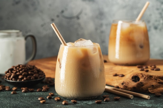 Ice coffee in a tall glass with cream poured over ice cubes and beans on a dark concrete table Cold summer drink with tubes on a black background with copy space