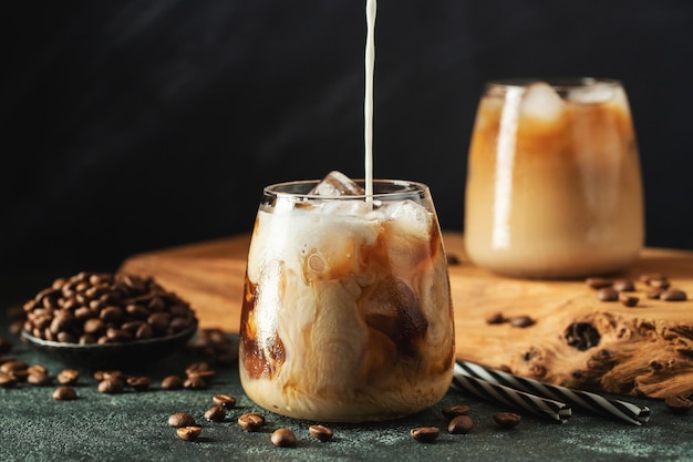 ice coffee in a tall glass with cream poured over ice cubes and beans on a dark concrete table cold summer drink with tubes on a black background with copy space