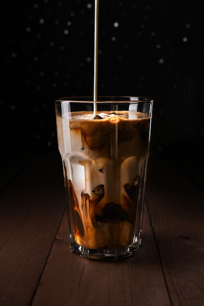 Ice coffee on a wood table with cream pouring into glass showing the texture and refreshing look of the drink, with a dark bokeh background.