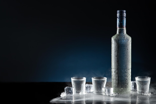 Photo ice-cold bottle of vodka with shot glasses on dark blue background with copy space.