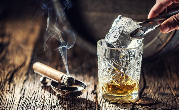 Ice cube being put into an ornamental cup of whisky placed on a old fashioned wood and burning cigar aside.