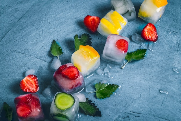 Foto cubetti di ghiaccio con frutta e ghiaccio rotto su una superficie di pietra blu con foglie di menta e frutta fresca. menta, fragola, ciliegia, limone, arancia. vista piana, vista dall'alto