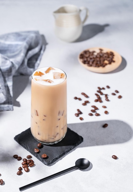 Iced coffee latte in a tall glass with milk on a light background with coffee beans milk jug and morning shadows Summer refreshment concept