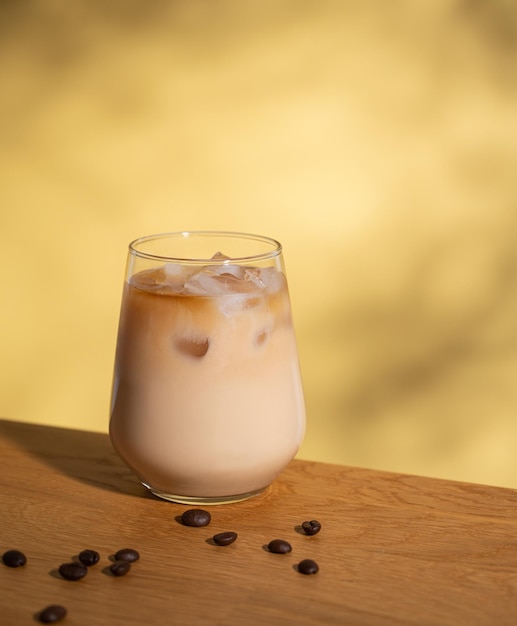 Iced coffee with milk in a glass with ice cubes and grains on a yellow background with shadows The concept of a cold summer drink Copy space