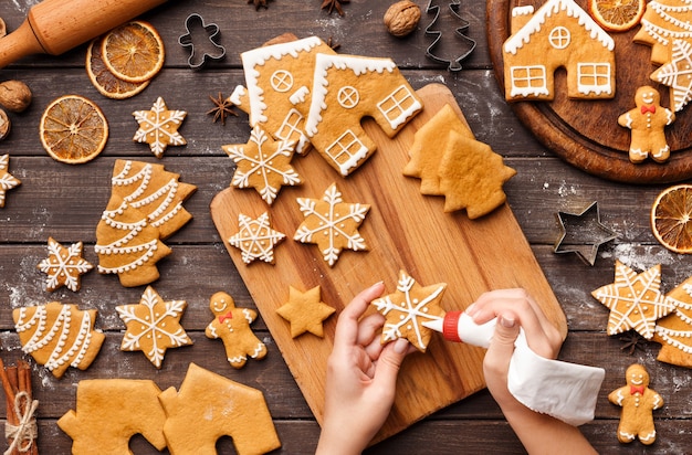 Icing process of Christmas bakery. Unrecognizable woman decorating homemade gingerbread cookies on wooden table, top view