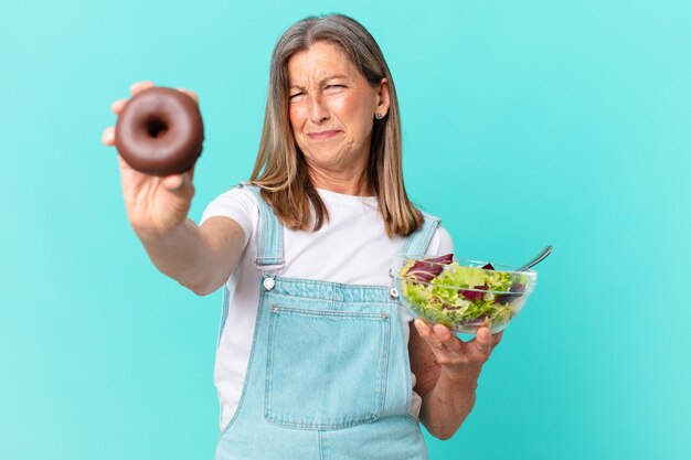 Donna graziosa di età inattiva con una ciambella e un concetto di dieta dell'insalata