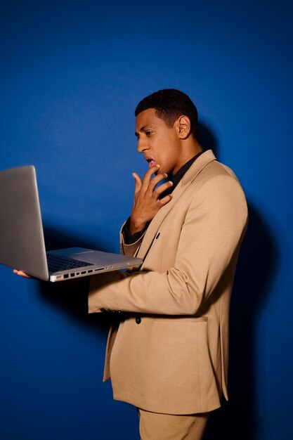 Idea. Dark-skinned guy in light stylish suit standing sideways to camera touching chin with his hand looking at laptop on blue background