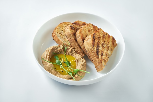 Идеальная закуска - паштет из печени индейки с соусом из манго и ржаным хлебом в белой тарелке на белом фоне