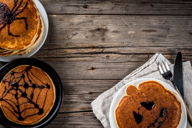 Идеи на завтрак Хэллоуин, еда для детей. Тыквенный пирог из блинов украшен шоколадным сиропом в традиционном стиле, паутиной, пауком, фонарем из тыквы. На деревянном деревенском столе,
