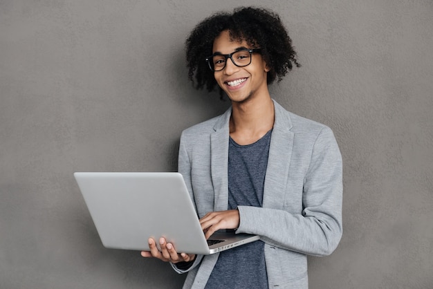 Ideeën laten gebeuren. Vrolijke jonge Afrikaanse man die laptop vasthoudt en glimlacht terwijl hij tegen een grijze achtergrond staat