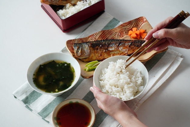 Iemand die een kom met gekookte rijst vasthoudt en eetstokjes gebruikt die rijst proberen te plukken geserveerd met een gegrilde saba of makreelvis en misosoep op witte en groene gestreepte placemat op witte tafel