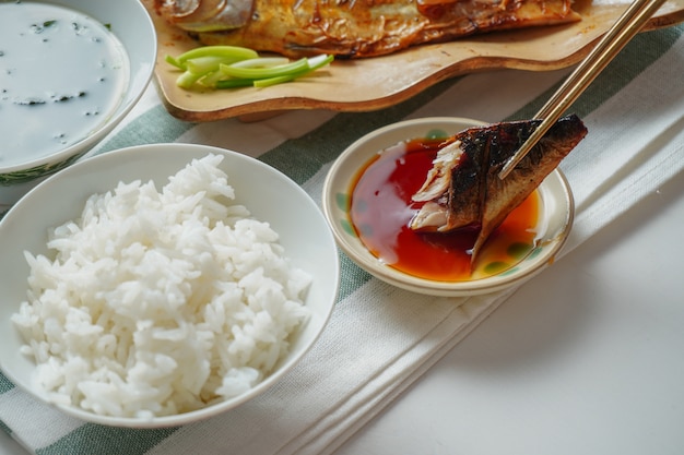 Iemand die eetstokjes gebruikt om een stuk gegrilde saba of makreelvis te plukken en op zoete saus te plakken, geserveerd met gekookte rijst en miso-soep op wit en groen gestreepte placemat op witte tafel