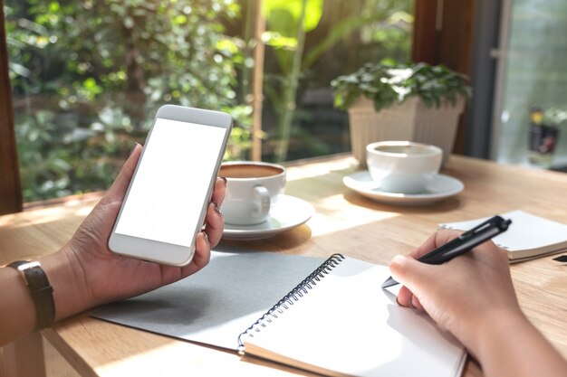 Iemand houdt een smartphone wit terwijl hij op een notebook schrijft