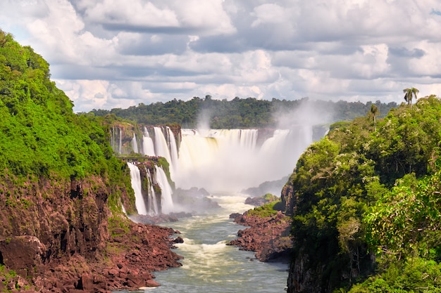 Cascate di iguazu in argentina. le imbarcazioni a motore turistiche vanno verso una potente cascata d'acqua creando nebbia sul fiume iguazu. lussureggiante fogliame della foresta pluviale subtropicale lungo le coste di pietra rossa.