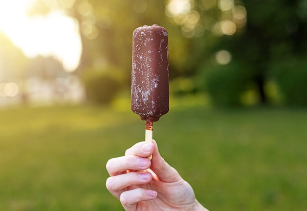 Ijs op stok in vrouwelijke hand close-up. Icecream op groene bomen op de achtergrond van de natuur.