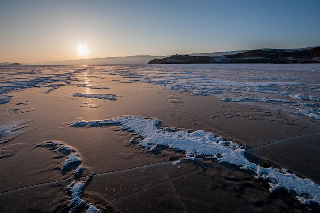Ijs van het Baikalmeer bij zonsondergang met scheuren en sneeuw Bergen voorbij het meer De zon is aan de horizon Weerspiegeling van de zon op ijs