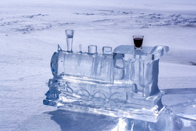 IJssculptuur in de vorm van een treinlocomotief en rode likeur in een glas. IJs lost op in de zon. Wazig sneeuw op de achtergrond. Horizontaal.