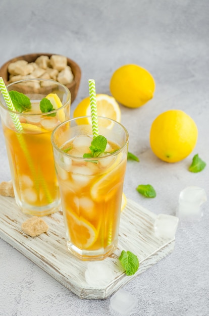 IJsthee met citroen, bruine suiker, muntblaadjes en ijsblokjes in een glas op een bord op een lichte achtergrond. Zomer verfrissend drankje.