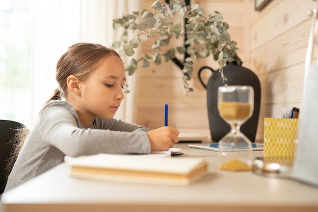 Ijverig schoolmeisje in grijze trui zittend aan tafel en pen vasthoudend over pagina van open notitieboekje terwijl ze huiswerk maakt bij bureau in slaapkamer