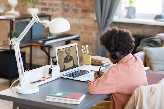 Ijverige schooljongen die aantekeningen maakt terwijl hij naar de leraar op het laptopscherm kijkt