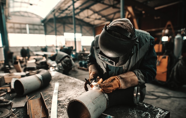 IJzerarbeider in beschermend pak, masker en handschoenen die pijp lassen. Werkplaats interieur.