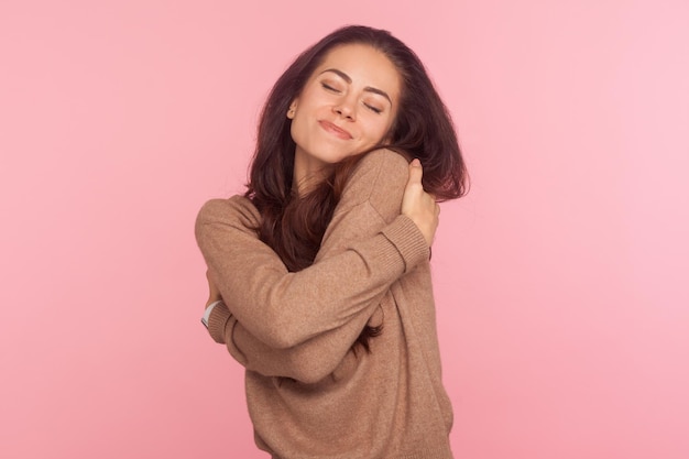 Ik ben egoïst, hou van mezelf. portret van een egoïstische narcistische jonge vrouw met donkerbruin haar die zichzelf omhelst en lacht met een dromerige tevreden uitdrukking. indoor studio-opname geïsoleerd op roze achtergrond