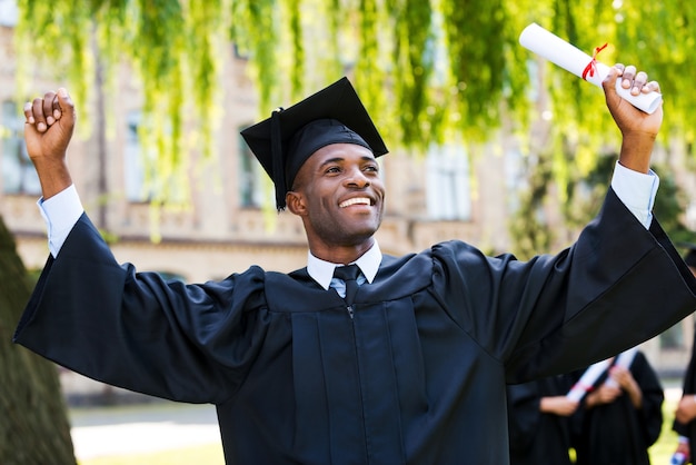 Ik ben eindelijk afgestudeerd! gelukkige jonge afrikaanse man in afstudeerjurken met diploma en armen omhoog terwijl zijn vrienden op de achtergrond staan