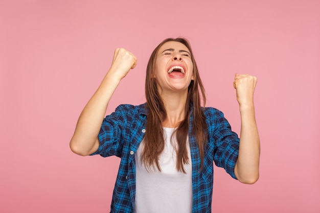 Ik ben kampioen portret van extatisch succesvol meisje in geruit hemd schreeuwen van vreugde met opgeheven vuisten vieren winnende ongelooflijke overwinning indoor studio opname geïsoleerd op roze achtergrond