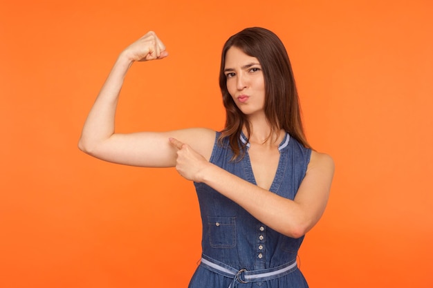 Ik ben sterk en onafhankelijk. Zelfverzekerde brunette vrouw die naar biceps wijst, trots naar de camera kijkt, kracht en kracht voelt om succes te behalen. indoor studio-opname geïsoleerd op oranje achtergrond