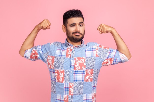 Ik ben sterk portret van een aantrekkelijke man in een casual blauw shirt die handen opsteekt om biceps te laten zien en zelfverzekerd naar de camera kijkt en kracht en kracht voelt Indoor studio shot geïsoleerd op roze achtergrond