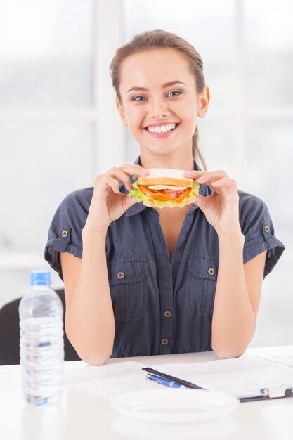 Ik heb wat pauze nodig. Mooie jonge vrouw die sandwich vasthoudt terwijl ze op haar werkplek zit