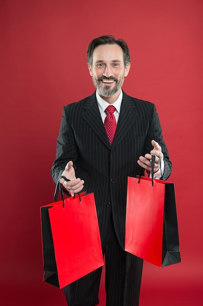 Ik help economie, ik winkel veel. Gelukkige shopper rode achtergrond. Zakenman houdt boodschappentassen vast. Volwassen man geniet van winkelen. Uitverkoop. Cadeautjes shoppen. Zwarte vrijdag. Goede dag om te winkelen.