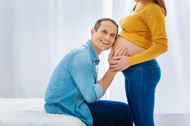 Ik hoor hem. Positief opgetogen toekomstige vader die een glimlach op zijn gezicht hield en handen op de buik van de baby legde terwijl hij vooruitkijkt