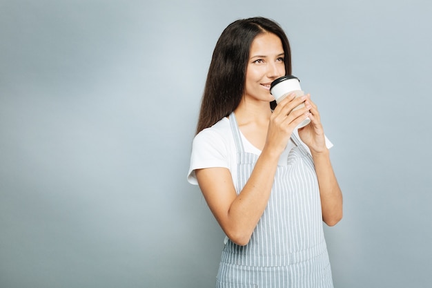 Ik hou van deze geur. Positieve brunette die positiviteit uitdrukt terwijl ze cappuccino drinkt en naar boven kijkt, geïsoleerd op grijs