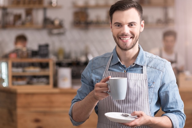 Ik hou van dit drankje. Knappe aangename jonge man glimlachend en kopje koffie te houden terwijl hij in een café.