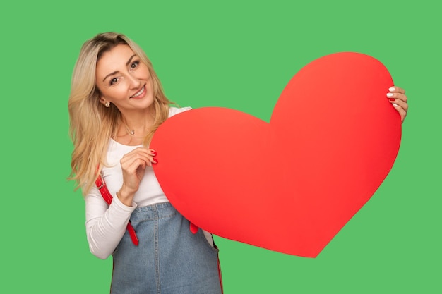 Ik hou van je, fijne Valentijnsdag. Portret van een aantrekkelijke volwassen blonde vrouw die een rood hart vasthoudt en speels glimlacht, flirt, romantische felicitatie. indoor studio-opname geïsoleerd op groene achtergrond