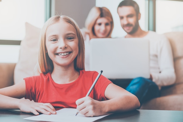 Ik hou van mijn familie! Gelukkig klein meisje dat iets op papier tekent en glimlacht terwijl haar ouders op de achtergrond zitten met laptop