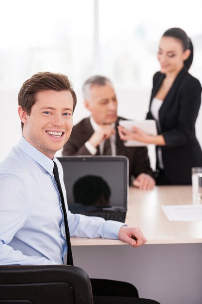 Ik hou van mijn werk. Knappe jonge man in formalwear die over de schouder kijkt en glimlacht terwijl hij aan tafel zit met twee andere zakenmensen die op de achtergrond praten